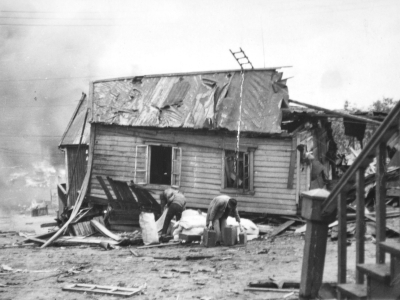 Svart-hvitt-bilde av et ødelagt hus. Taket er rasert og vi ser røyk i bakgrunnen. Foto